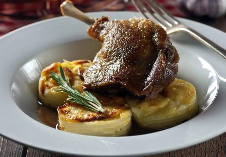 Un plato blanco con confit de pato con patatas dauphinoise y una ramita de romero fresco